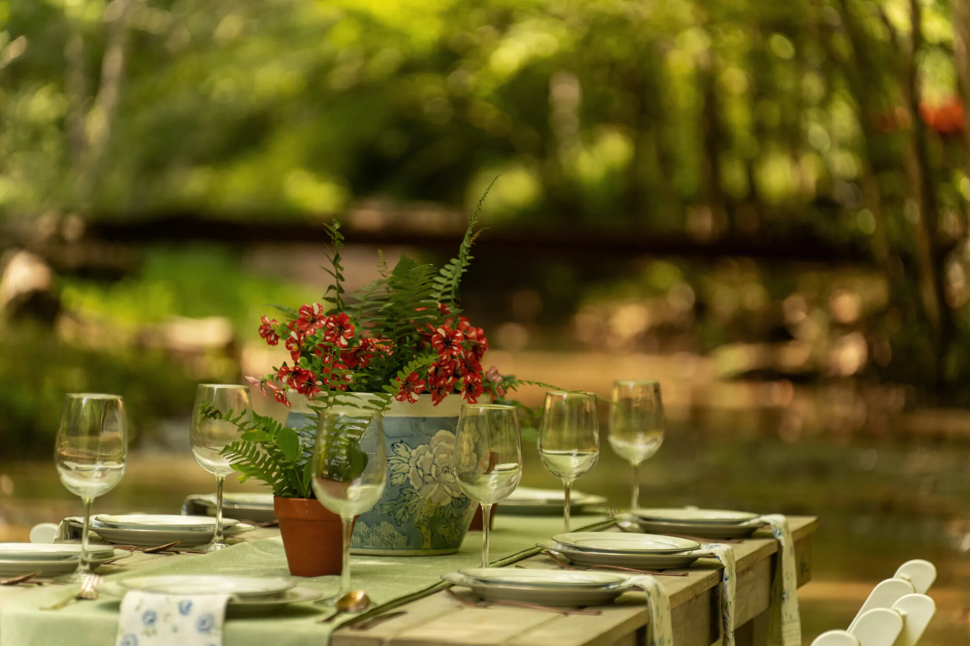 A blue-and-white planter of red blooms and ferns at the centre of the table, the log footbridge blurred beyond