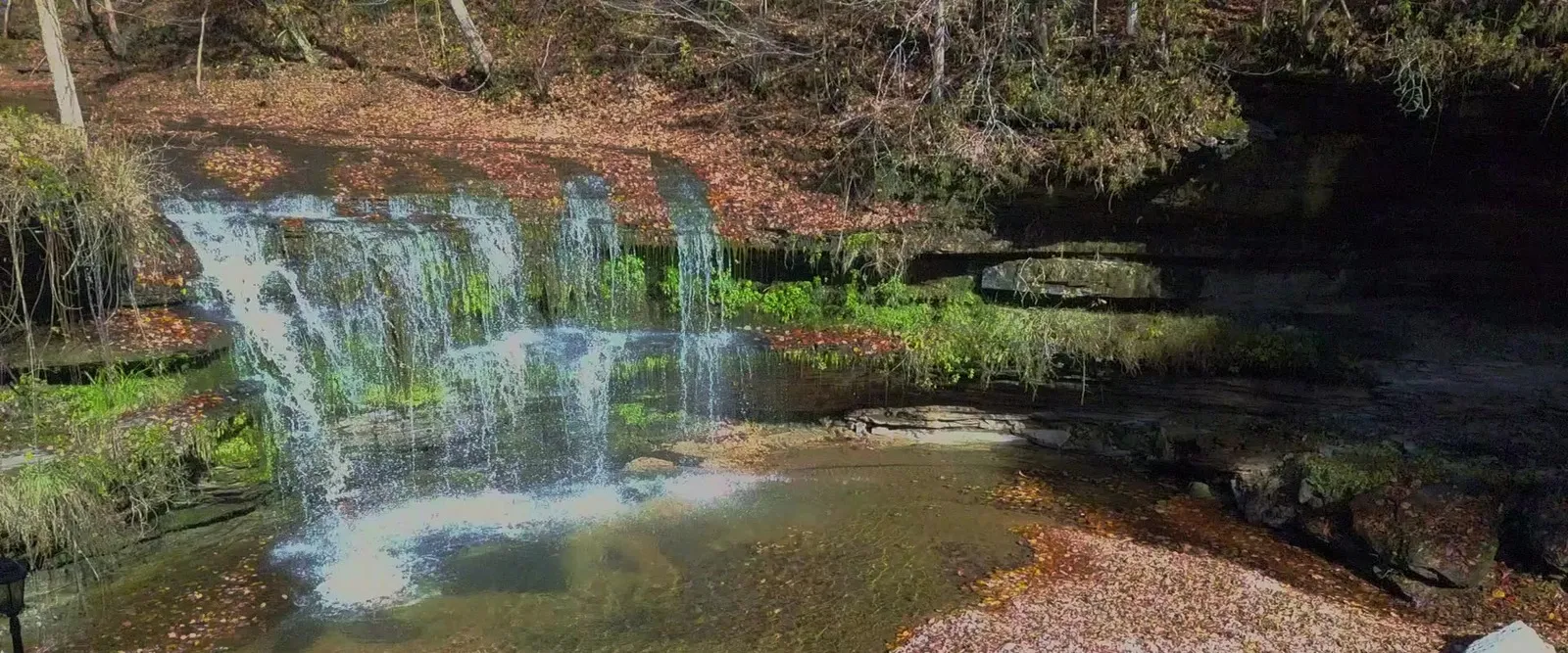 Aerial view of Old Falls pouring over the limestone shelf into its sunlit plunge pool, ringed by autumn leaves