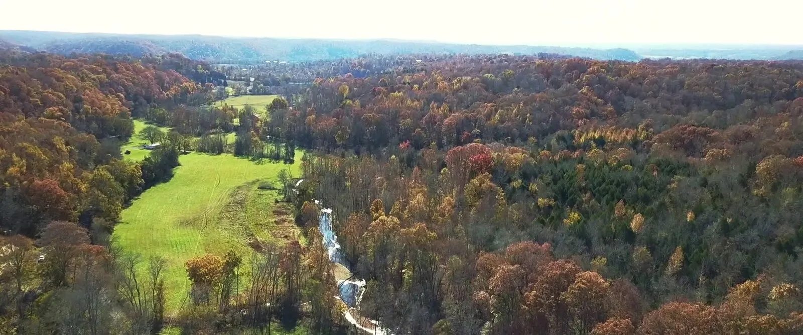 Aerial view of the wooded hollow with green pasture, autumn ridges, and the creek winding down toward the falls