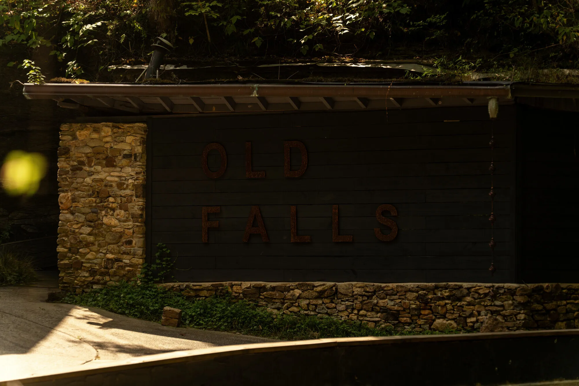 The OLD FALLS sign wall in deep afternoon shade, its stone base and rusted letters catching the last light