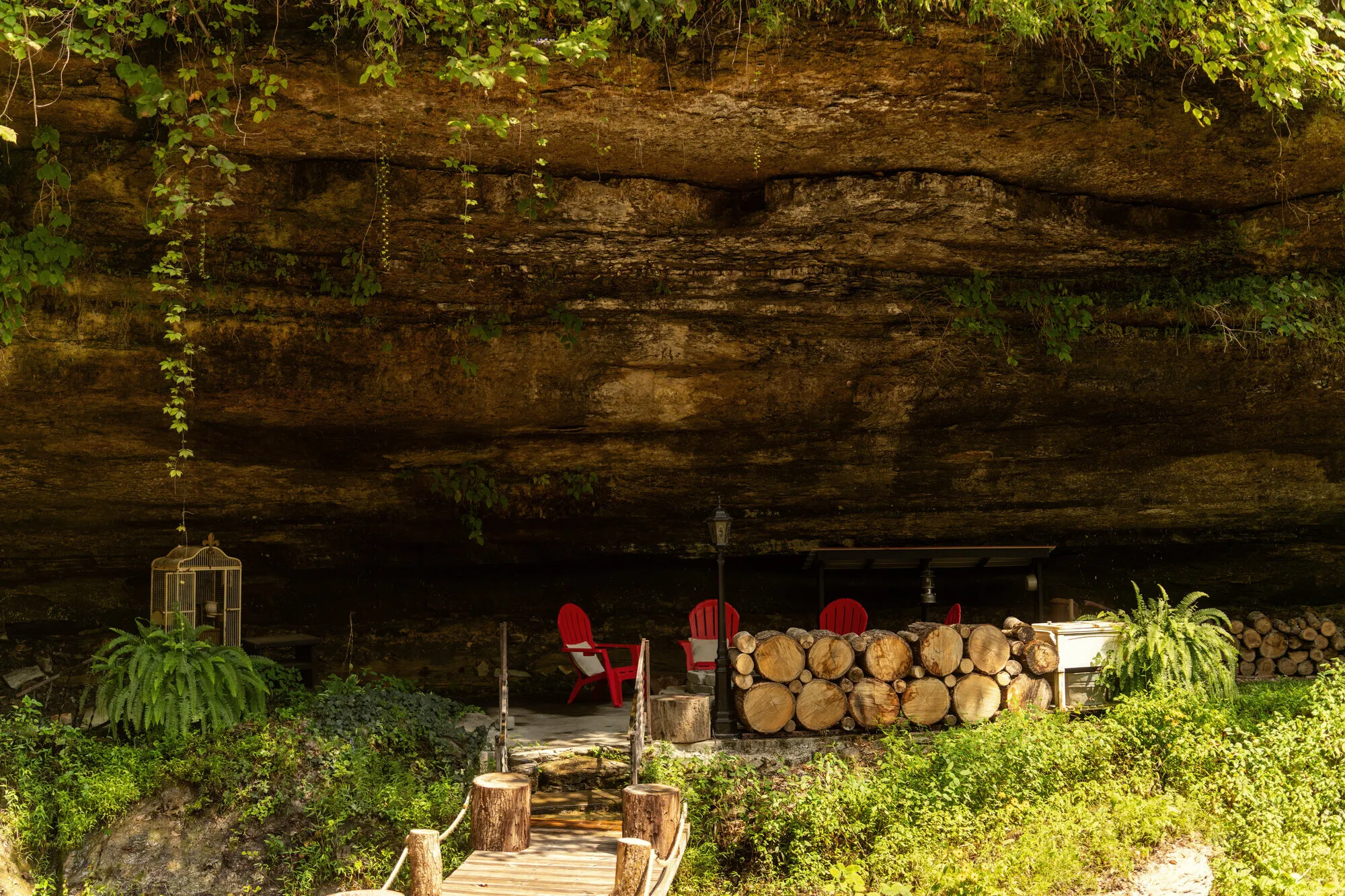 Red Adirondack chairs and a stacked wall of cut firewood sheltering under the overhanging limestone ledge, a plank walkway leading in