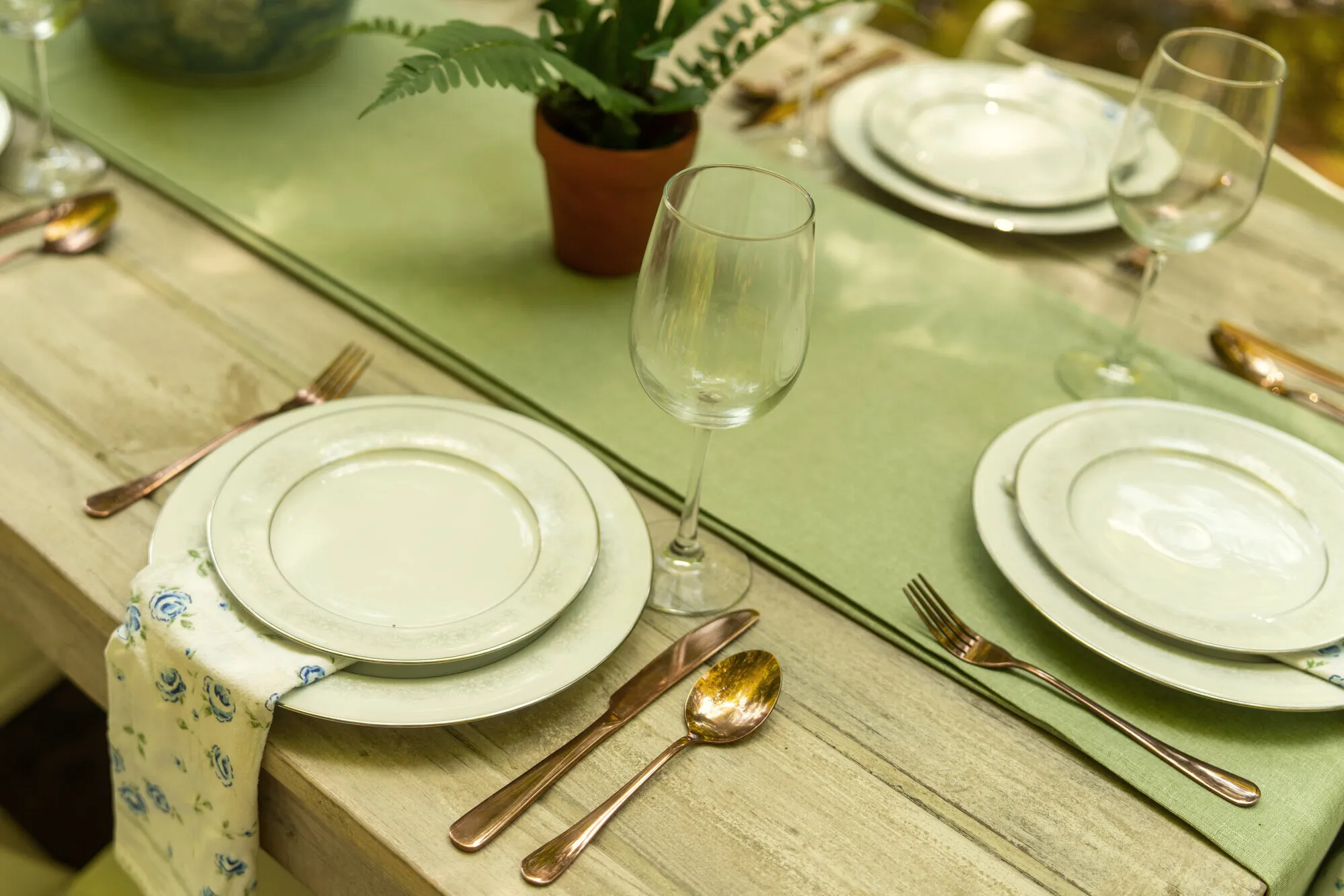A place setting of gold-rimmed china and copper cutlery on a pale green runner, a blue-rose napkin folded alongside