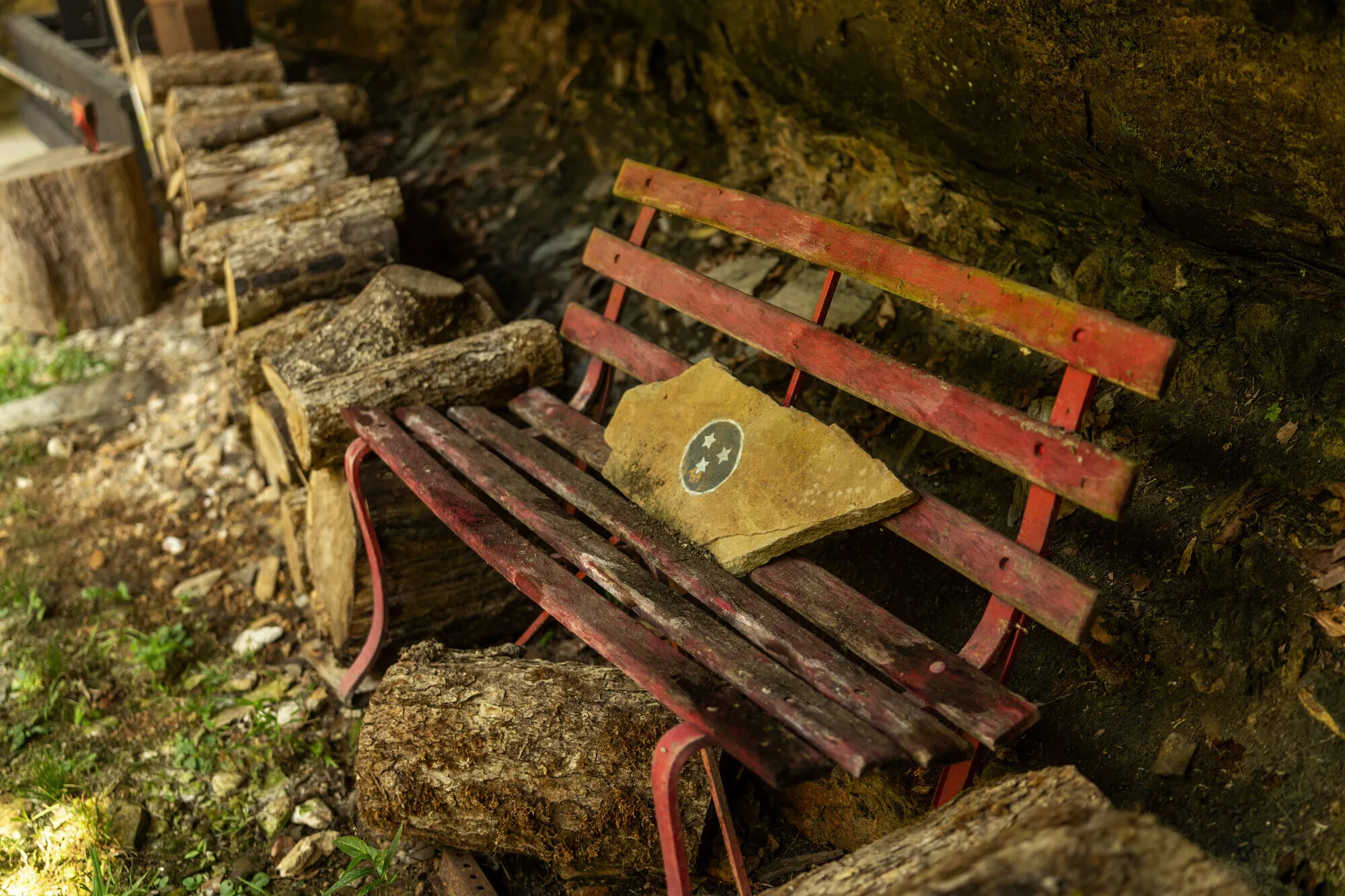 A weathered red garden bench under the rock ledge, a Tennessee tri-star painted on a slab of creek stone resting on its seat